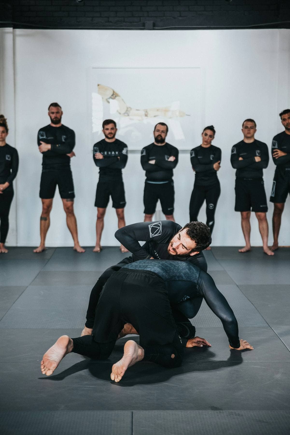 Trainer zeigt eine Technik am Boden, dahinter steht das Team in gleichen Rashguards